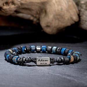 Könnte beinhalten: Ein Armband mit einer Mischung aus schwarzen, blauen und silbernen Perlen. Es hat einen rechteckigen Silberanhänger mit dem eingravierten Namen "Adam" und dem Datum "14.7.24". Die Perlen sind auf einer schwarzen Schnur aufgefädelt.