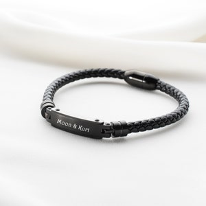 May include: Black leather bracelet with a black metal clasp and a rectangular plate engraved with the text "Moon & Kurt".