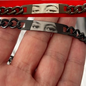 May include: A black metal chain bracelet featuring a rectangular central plate. The plate displays a detailed black and white graphic print of a pair of human eyes with dark eyeliner, integrated into the smooth, dark metallic surface.