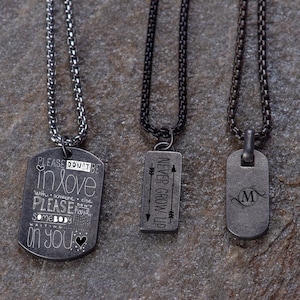 May include: Three silver chain necklaces with different pendants. The first pendant is a silver dog tag with the text "PLEASE DON'T BE IN LOVE WITH SOMEONE ELSE PLEASE DON'T HAVE SOMEBODY WAITING ON YOU". The second pendant is a silver rectangle with the text "NEVER GROW UP" and two arrows. The third pendant is a silver dog tag with the letter "M" in a decorative font.