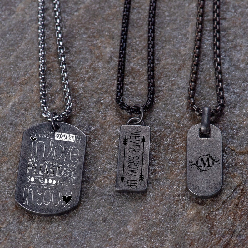 May include: Three silver chain necklaces with different pendants. The first pendant is a silver dog tag with the text "PLEASE DON'T BE IN LOVE WITH SOMEONE ELSE PLEASE DON'T HAVE SOMEBODY WAITING ON YOU". The second pendant is a silver rectangle with the text "NEVER GROW UP" and two arrows. The third pendant is a silver dog tag with the letter "M" in a decorative font.
