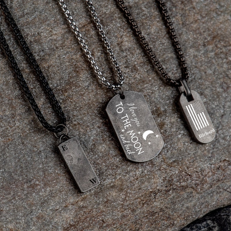 May include: Three silver chain necklaces with different pendants. The first pendant is a rectangular tag with the text "I love you to the moon and back" and a crescent moon and stars. The second pendant is a rectangular tag with the letters "E" and "W". The third pendant is a rectangular tag with an American flag and the name "Richard".