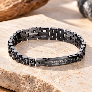 May include: A black metal link bracelet featuring a rectangular plate engraved with the coordinates 34°41'21.4"N 48°67'54.9"W and the Roman numeral date VII.XXVI.MMXXIV. The bracelet has a multi-link chain design with a matte finish.