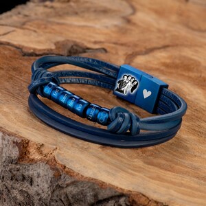 May include: A blue leather bracelet with a photo charm and engraved beads. The beads are engraved with the names Eric, John, Nina, Eva, and the date 13.10.20. The bracelet also has an infinity symbol charm.