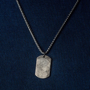 May include: A silver chain necklace with a silver dog tag pendant. The dog tag is blank and has a slightly worn look.