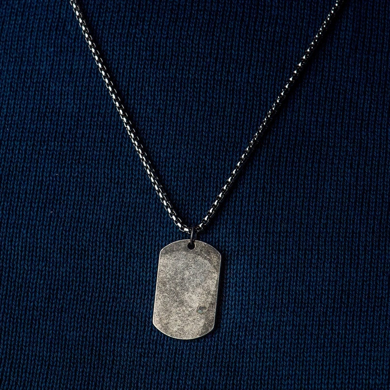 May include: A silver chain necklace with a silver dog tag pendant. The dog tag is blank and has a slightly worn look.