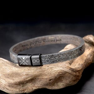 Puede incluir: Una pulsera de cuero gris con un cierre negro. La pulsera tiene el texto grabado "I love you to the moon and back". El cierre presenta las iniciales "W M" con un corazón, el número "99" y un símbolo de infinito.