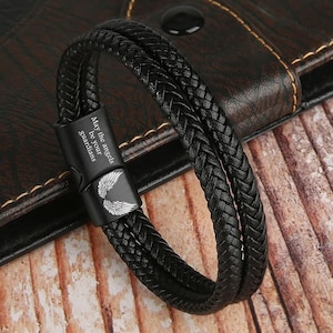 May include: Black braided leather bracelet with a magnetic clasp. The clasp features the text "May the angels be your guardians" and a wing design. The bracelet is shown on a wooden surface.