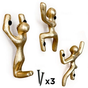 May include: Three gold-colored metal hooks shaped like stylized figures in a climbing pose. The hooks are mounted on a white wall. The hooks come with three black screws.  The text "x3" is printed below the screws.