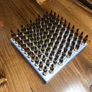May include: A silver ammunition tray filled with many rifle bullets. The bullets have brass casings and pointed tips. The tray is on a wooden surface with a visible wood grain. The bullets are arranged in a grid.