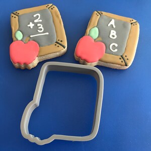 Könnte beinhalten: Schul-Themen-Kekse. Zwei Kekse in Form von Tafeln mit den Zahlen 2 + 3 und den Buchstaben A, B, C. Jeder Keks hat einen roten Apfel mit einem grünen Blatt. Ein grauer Ausstecher befindet sich im Vordergrund.