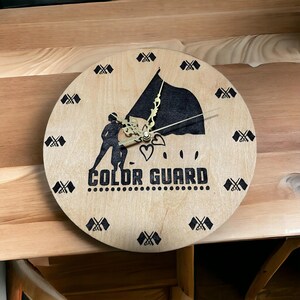 May include: A wooden clock with a black and white design of a color guard holding a flag. The clock face has the words "COLOR GUARD" and is surrounded by small black and white flag designs.