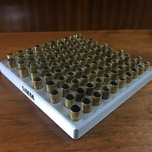 May include: A grey 9mm ammunition loading block filled with brass casings. The block has multiple holes to hold the casings upright. The text "9MM" is printed on the side. The casings are a shiny gold colour.