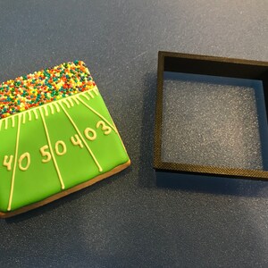 Pode incluir: Um biscoito quadrado decorado como um campo de futebol americano com glacê verde, linhas brancas e granulado colorido. Um cortador de biscoito quadrado preto está ao lado do biscoito. O biscoito tem os números 0, 3, 40, 50 e 40.