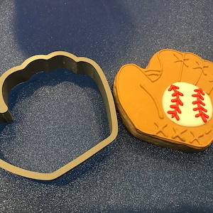 May include: A baseball-themed cookie cutter next to a baseball glove-shaped cookie. The cookie is tan with a white circle and red stitching, resembling a baseball. The cutter is gray.