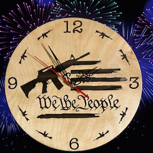 May include: A round wooden clock with black numerals and hands. The clock face features a rifle design, the words "We the People", and small rifle silhouettes. The second hand is red.
