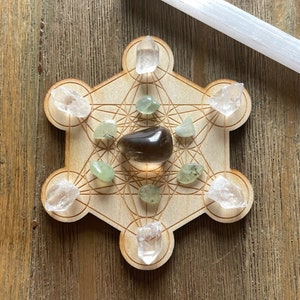 May include: A wooden grid with a geometric pattern, known as a sacred geometry grid, with clear quartz crystals and green aventurine crystals placed on the points. A smoky quartz crystal is placed in the center of the grid.