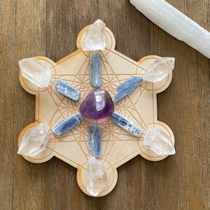Peut inclure: Une grille en bois du cube de Métatron avec une améthyste, des cristaux de kyanite bleue et de quartz clair disposés dessus. Une baguette de sélénite blanche repose à proximité. L'arrangement est sur une surface en bois.