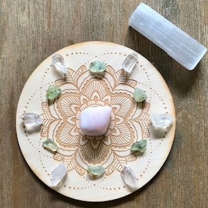 以下が含まれることがあります： 彫刻されたデザインと中央に配置された8つのクリスタルを持つ木製のマンダラ。クリスタルは透明、緑、ピンクです。画像には白いセレナイトスティックも含まれています。