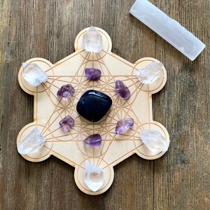 Puede incluir: Una rejilla de madera con un patr&oacute;n geom&eacute;trico, conocida como rejilla de geometr&iacute;a sagrada, con siete cristales de cuarzo transparente y siete cristales de amatista colocados en los puntos. Un cristal de obsidiana negra se coloca en el centro de la rejilla. Una varita de selenita blanca se coloca sobre una superficie de madera junto a la rejilla.