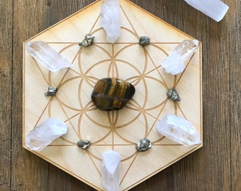 May include: A wooden hexagon-shaped crystal grid with a flower of life design. The grid has seven clear quartz crystals, six small pyrite crystals, and a tiger's eye stone in the center. A clear selenite wand is also included.