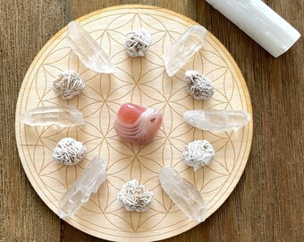 May include: A wooden circular tray with a flower of life design, featuring a pink agate crystal, seven clear quartz crystals, and seven selenite crystals.