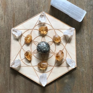May include: A wooden hexagon-shaped crystal grid with a pyrite crystal in the center and eight citrine crystals surrounding it. There are six clear quartz crystals around the perimeter of the grid. A clear quartz crystal wand is also included.