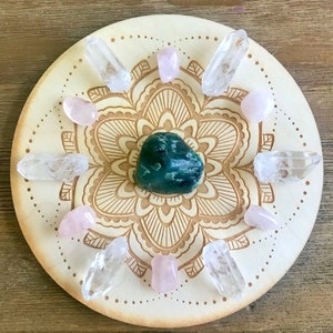 May include: A wooden crystal grid with a mandala design. The grid features a green aventurine crystal in the center, surrounded by clear quartz crystals and rose quartz crystals.