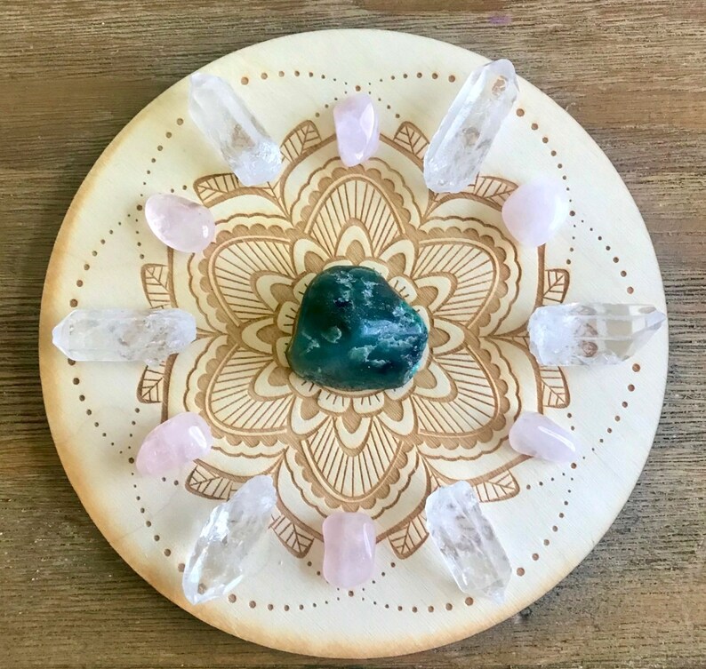 May include: A wooden crystal grid with a mandala design. The grid features a green aventurine crystal in the center, surrounded by clear quartz crystals and rose quartz crystals.
