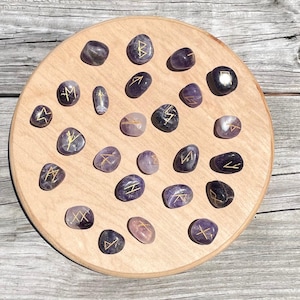 May include: A collection of twenty-four polished, purple amethyst stones arranged on a circular light-colored wooden board. Each stone features a unique, hand-engraved golden rune symbol etched into its smooth, rounded surface.