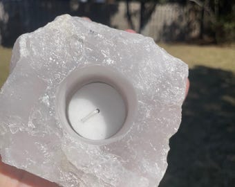 May include: A rough-hewn, light pink quartz crystal candle holder. The natural stone has a carved-out circular space to hold a small white tea light candle. The candle holder is held in a hand against a blurred outdoor background.
