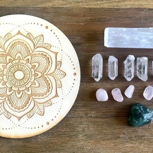May include: A wooden mandala with a carved floral design, surrounded by various crystals including clear quartz points, rose quartz tumbles, a selenite stick, and a green aventurine stone.