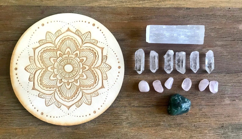 May include: A wooden mandala with a carved floral design, surrounded by various crystals including clear quartz points, rose quartz tumbles, a selenite stick, and a green aventurine stone.