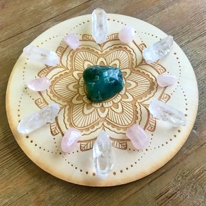 May include: A wooden mandala crystal grid with a green crystal in the center, surrounded by clear and pink crystals.