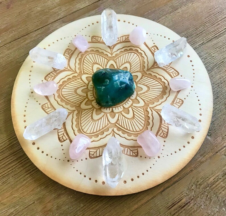 May include: A wooden mandala crystal grid with a green crystal in the center, surrounded by clear and pink crystals.