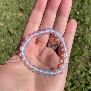 May include: A bracelet made of iridescent opal beads. The beads are a light purple color with a white center. The bracelet is on a person's hand.