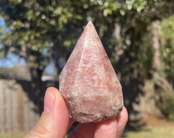 May include: A faceted, pinkish-orange crystal point held in a hand. The crystal has a rough, natural base and a smooth, polished top. The background is blurred, showing green foliage and sunlight.