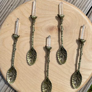 May include: Five ornate gold spoons with clear quartz crystal handles. The spoons have a floral design and are arranged on a wooden surface.