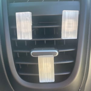 May include: Three clear plastic air freshener clips hanging from the air vents in a car. The air freshener clips are hanging from the center vent and the two side vents.