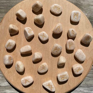 May include: A collection of twenty-four beige rune stones with gold-colored etched symbols, arranged on a round, light brown wooden board. The stones vary in shape and size, each displaying a unique runic character. The board sits on a wooden surface.