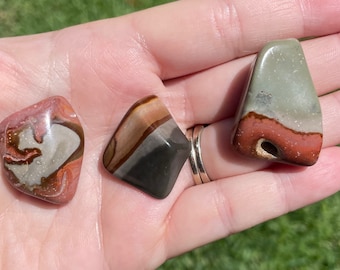 May include: Three smooth, polished stones with unique patterns of brown, red, and green. The stones are shaped like diamonds and have a natural, earthy look.