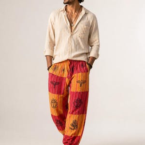 Puede incluir: Una persona con una camisa crema abotonada y pantalones harén estampados. Los pantalones presentan paneles rojos y naranjas con diseños gráficos negros. La imagen está sobre un fondo blanco liso.