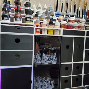 May include: A white and black modular storage unit with multiple drawers and shelves. The unit is filled with various bottles of paint, brushes, and miniature figures. The unit has a glass door with a shelf for displaying miniature figures.