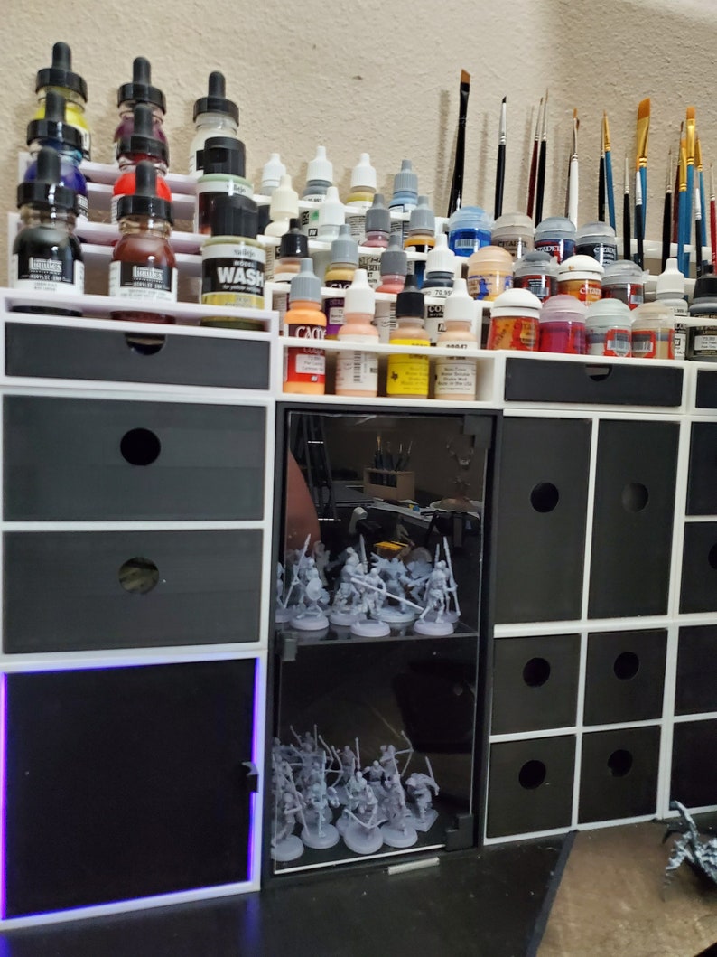 May include: A white and black modular storage unit with multiple drawers and shelves. The unit is filled with various bottles of paint, brushes, and miniature figures. The unit has a glass door with a shelf for displaying miniature figures.