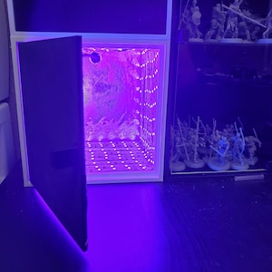 May include: A white cube with a purple light inside and a black door that is open. The cube is sitting on a black surface.