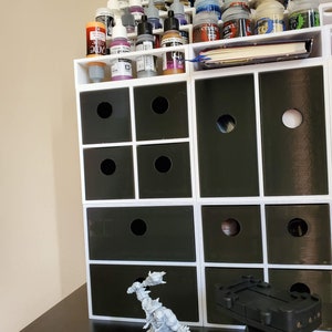 May include: A white organizer with multiple drawers, filled with small bottles of paint. The organizer is on a black surface with a gray plastic miniature figure of an orc holding a weapon.