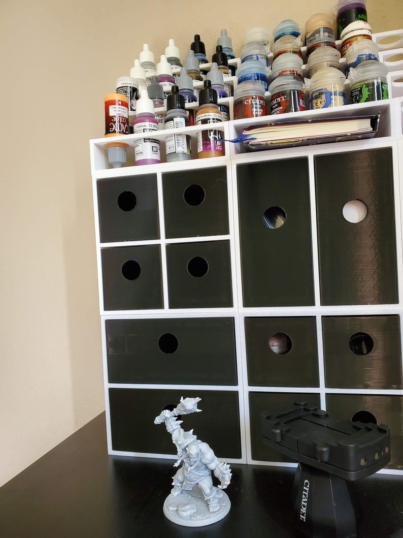 May include: A white organizer with multiple drawers, filled with small bottles of paint. The organizer is on a black surface with a gray plastic miniature figure of an orc holding a weapon.