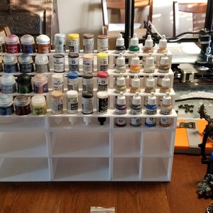 May include: White plastic paint rack with multiple rows of small paint bottles. The bottles are filled with various colors of paint. The rack is sitting on a wooden table.
