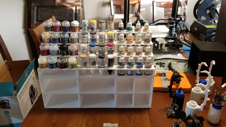 May include: White plastic paint rack with multiple rows of small paint bottles. The bottles are filled with various colors of paint. The rack is sitting on a wooden table.
