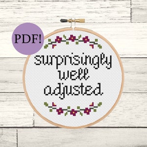 May include: A cross-stitch embroidery hoop with the words "surprisingly well adjusted" in black. The design includes a floral border with green leaves and purple flowers. A purple circle with "PDF!" is in the upper left corner.